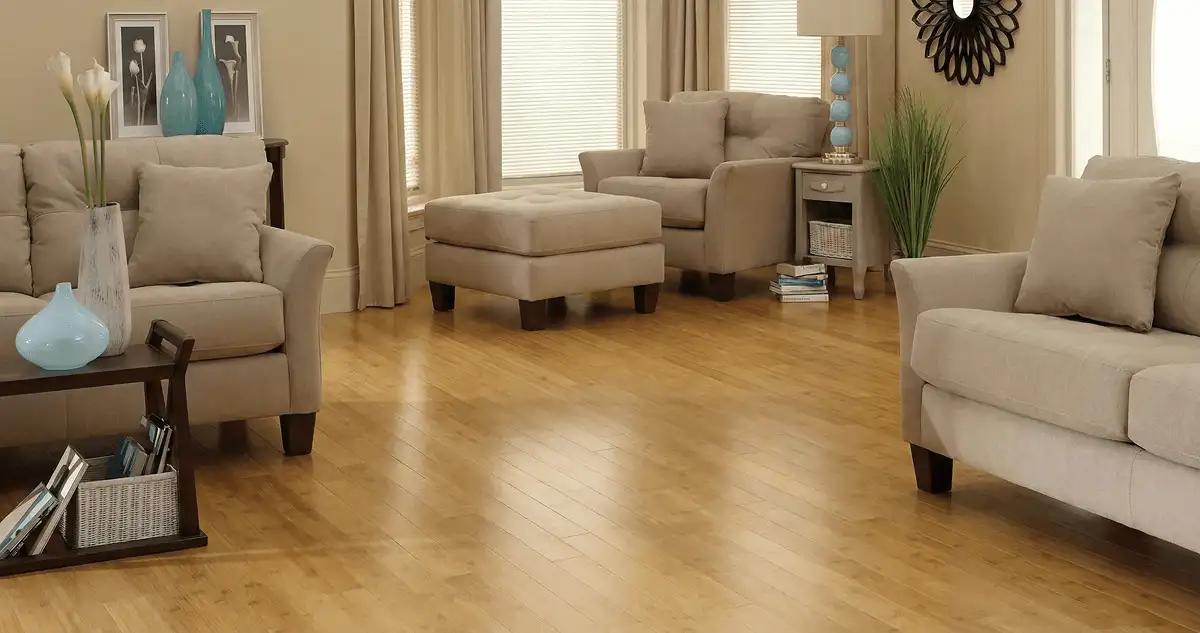 A living room with professionally installed bamboo flooring, beige sofas, a matching ottoman, a side table with a blue lamp, vases, framed pictures, and large windows with blinds and curtains in Fort Collins, CO